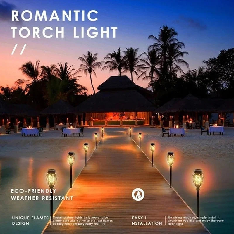 A wooden pathway lined with glowing torch lights leading toward a tropical silhouette at sunset.