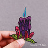 A hand holds an embroidered patch featuring a purple candle with a blue flame and red floral leaves.