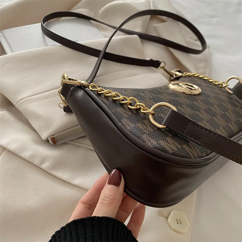 A dark brown PU leather crossbody bag with a gold chain strap is held by a hand against a light beige fabric background.