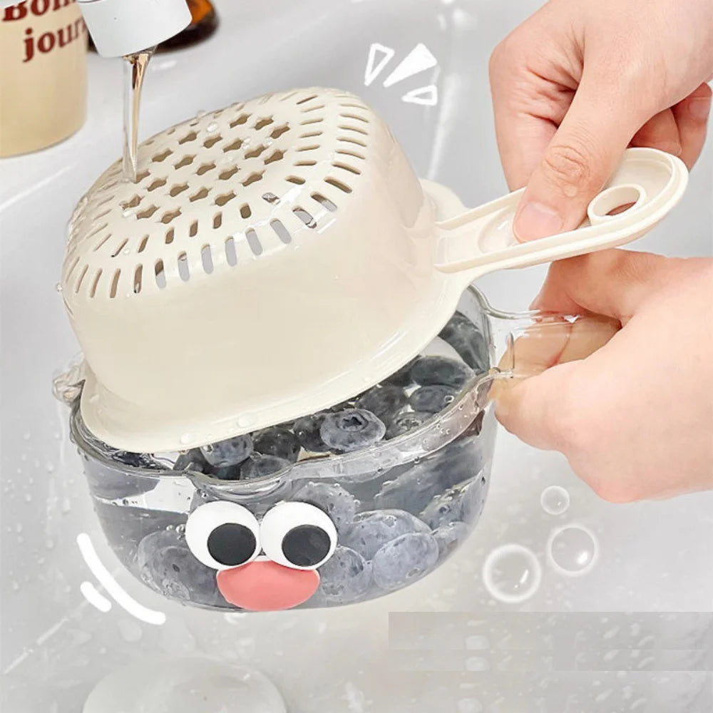 A hand is pouring liquid into a double-layer fruit washing basket with a perforated lid and a small face decoration.