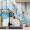 A black kitchen faucet is installed on a marble-patterned wall featuring blue and gold veining, with white utensils hanging a