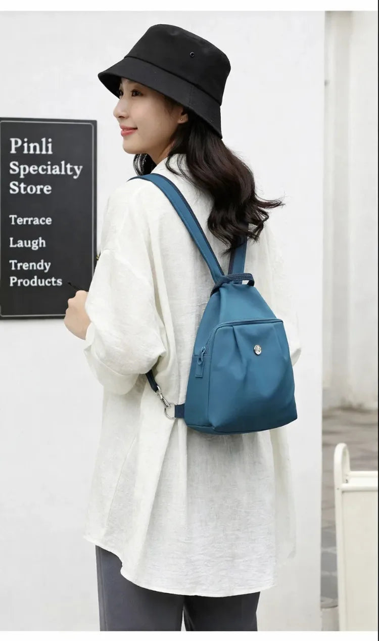 A woman wearing a white oversized shirt and black hat carries a teal mini backpack with a strap.