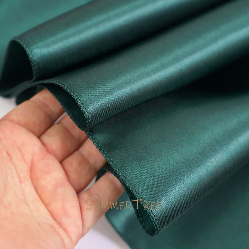 A hand is holding a piece of dark emerald green satin fabric, showing its smooth texture and drape.