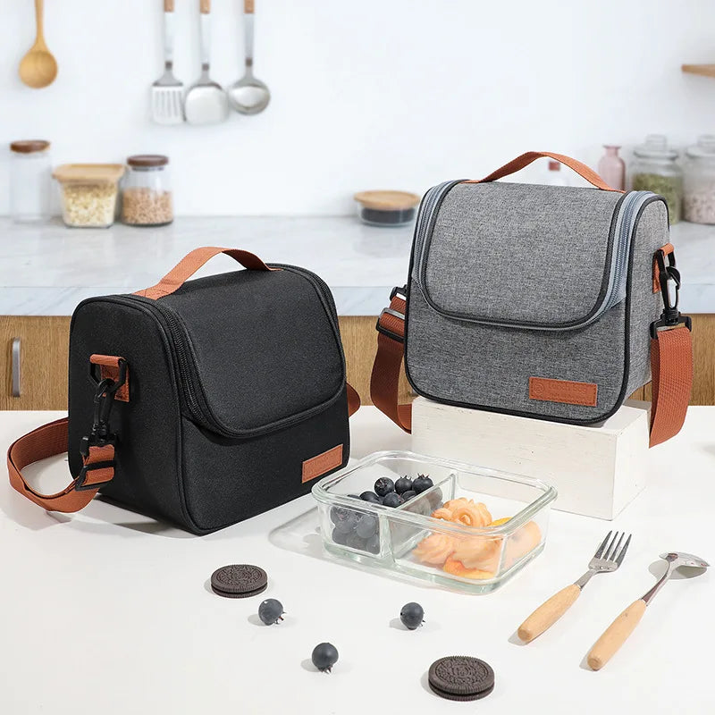 A gray insulated lunch bag with brown straps rests on a white table next to various kitchen utensils and food items.