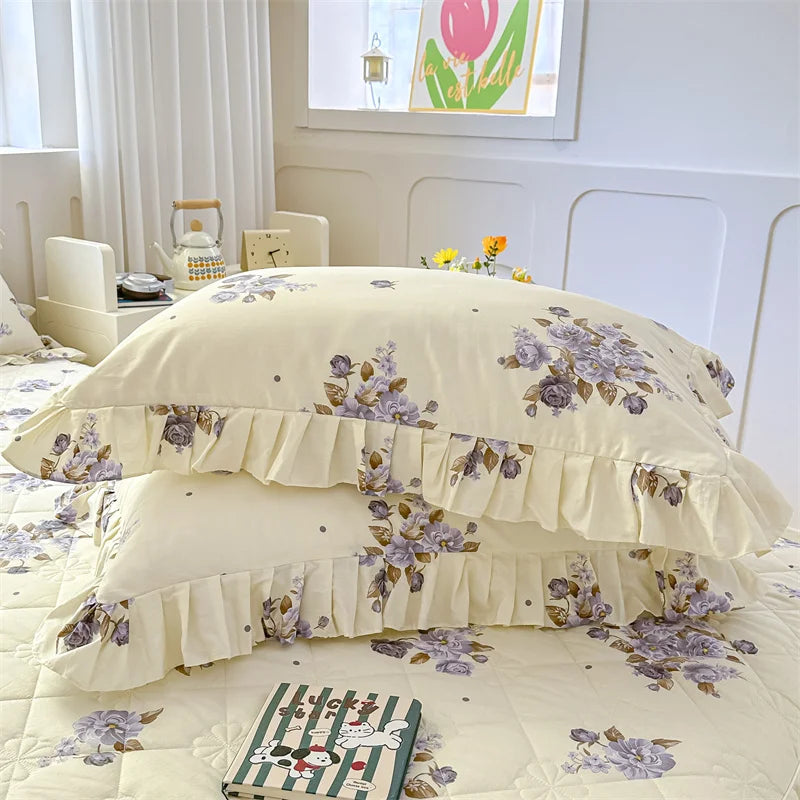 A set of two floral cotton pillowcases in pale yellow with purple and green floral patterns on a bed.