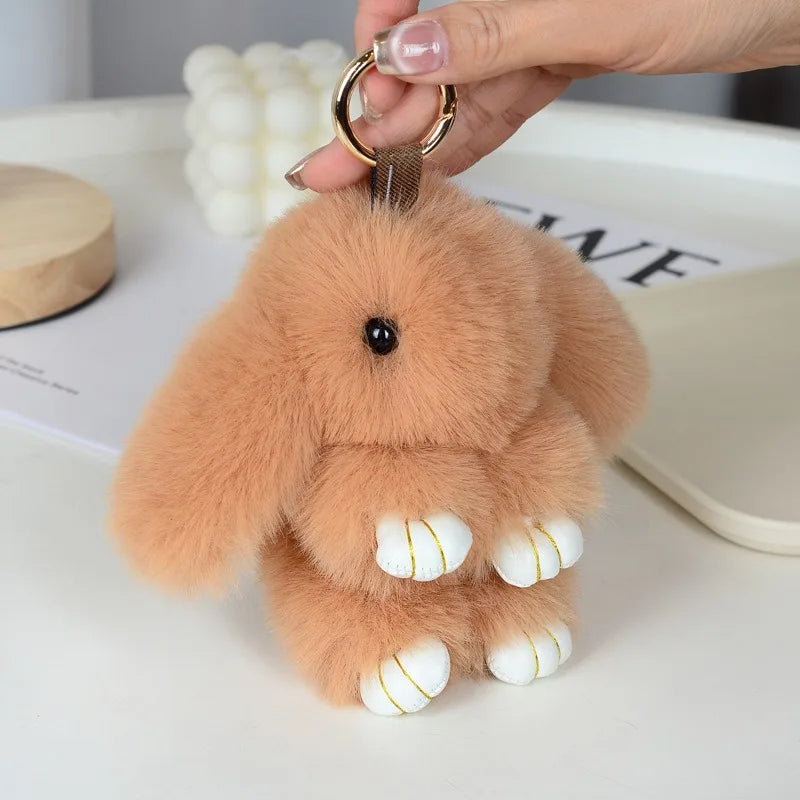 A fluffy, light brown bunny keychain with a pom pom tail is being held by a hand.