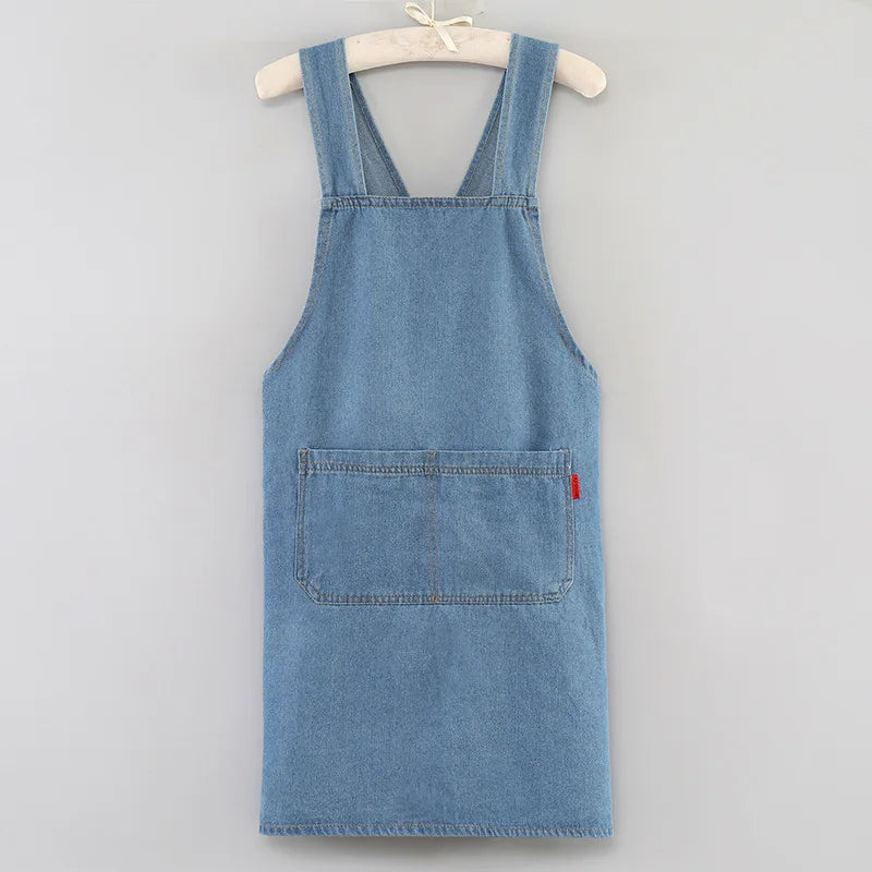 A light blue denim cross-back apron with chest pockets and a small red tag hanging from the pocket.