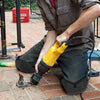 A person wearing dark clothing is using a yellow power tool on a brick floor while wearing black non-slip work knee pads.