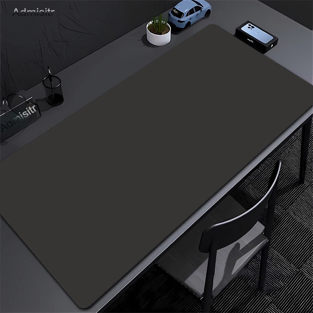 A dark gray oversized non-slip desk mat is shown on a desk with a laptop, a potted plant, and a smartphone.