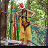 A child wearing a helmet is positioned on a wooden beam, holding a rope sling for climbing practice in an outdoor setting.