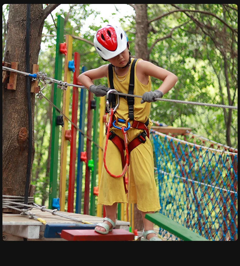 A child wearing a helmet is positioned on a wooden beam, holding a rope sling for climbing practice in an outdoor setting.