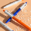 Three plastic pens in orange, white, and blue are shown spraying water on a light background.