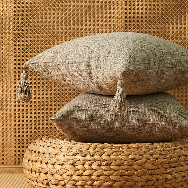 Two beige cotton linen throw pillows with tassels resting on a woven natural fiber surface against a textured wall.