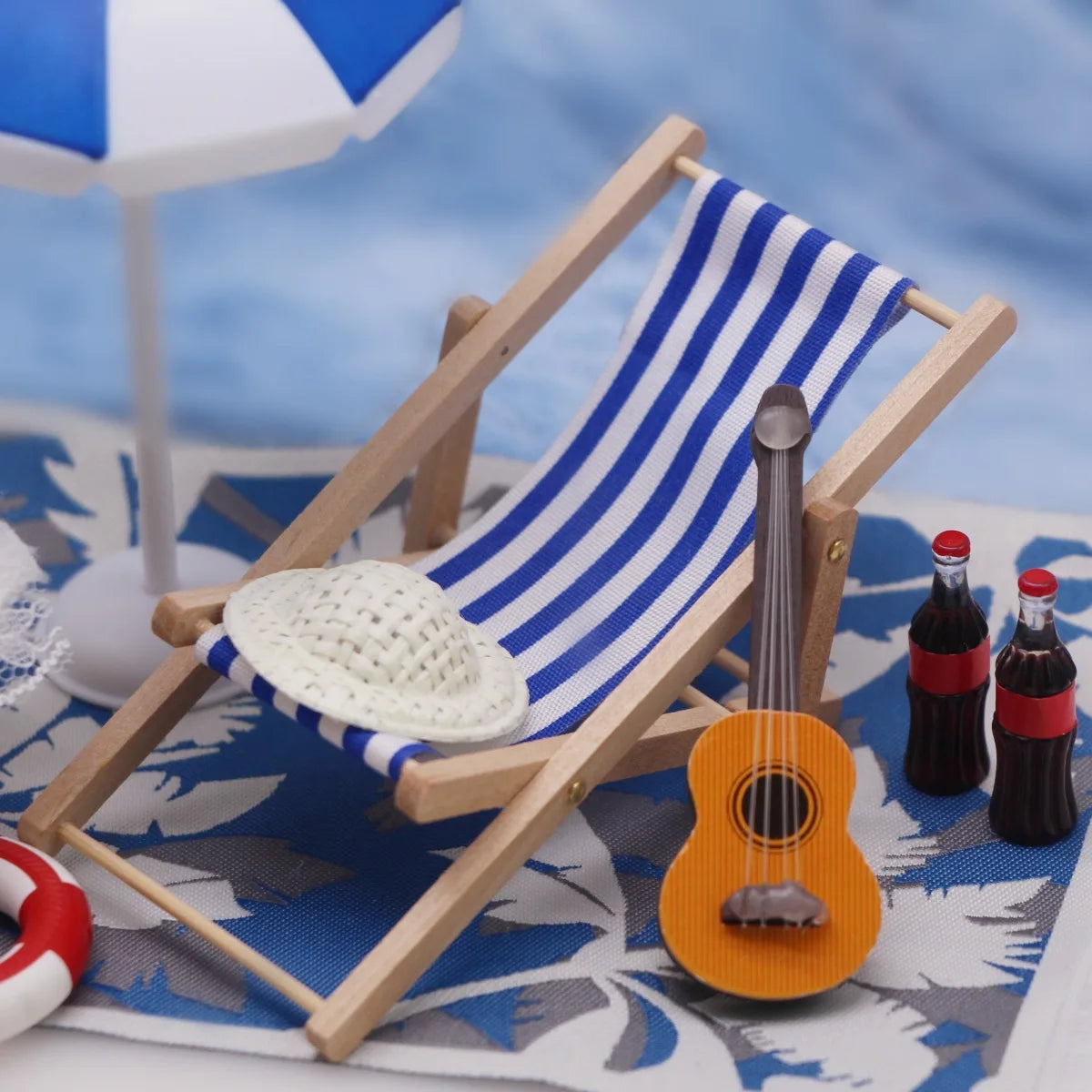 A wooden miniature beach chair with a striped towel and an orange ukulele-shaped bottle set on a blue cloth.