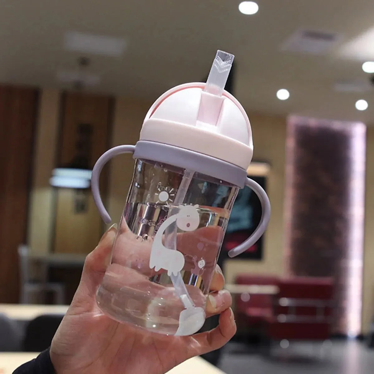 A hand holds a clear plastic sippy cup with a pink lid and gravity straw, featuring a white silhouette of a giraffe.