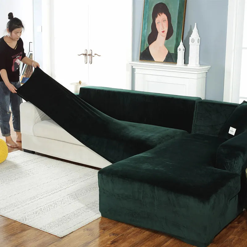 A dark emerald green plush velvet elastic sofa cover is shown draped over an L-shaped sofa in a brightly lit room.