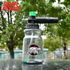 A clear glass bottle filled with liquid, featuring a black foam cannon attachment and a green nozzle, set against an outdoor 