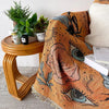 A bohemian jacquard woven throw blanket in earthy orange tones with stylized eye patterns draped next to potted greenery.