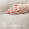 A hand gently touches a natural linen fringed pillow cover with a soft, neutral beige texture.