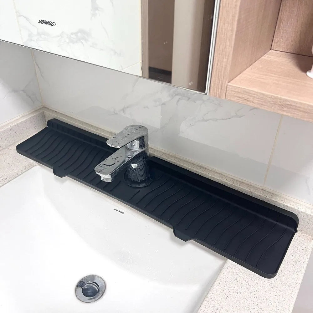 A black silicone faucet splash mat with a chrome faucet is placed on a white countertop next to a sink.
