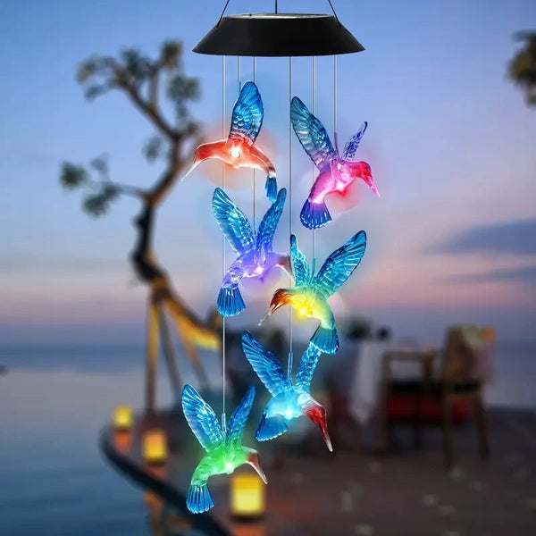 A hanging light fixture featuring multiple illuminated hummingbird-shaped glass or acrylic figures against a twilight sky.