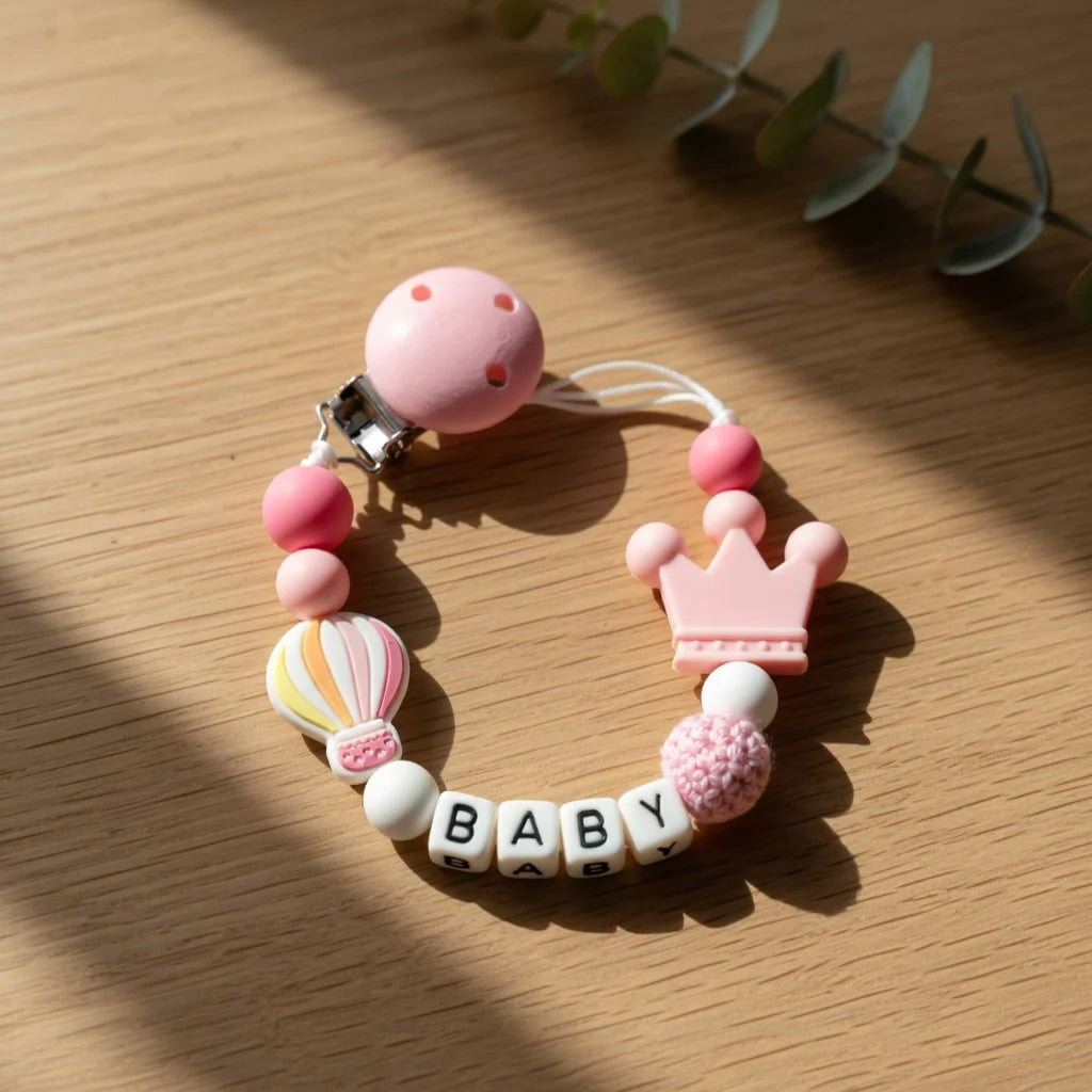 A pink baby pacifier clip with a crown charm and a beaded bracelet spelling out BABY.