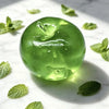 A smooth, translucent green fruit-shaped squeeze toy rests on a white marble surface with fresh mint leaves.