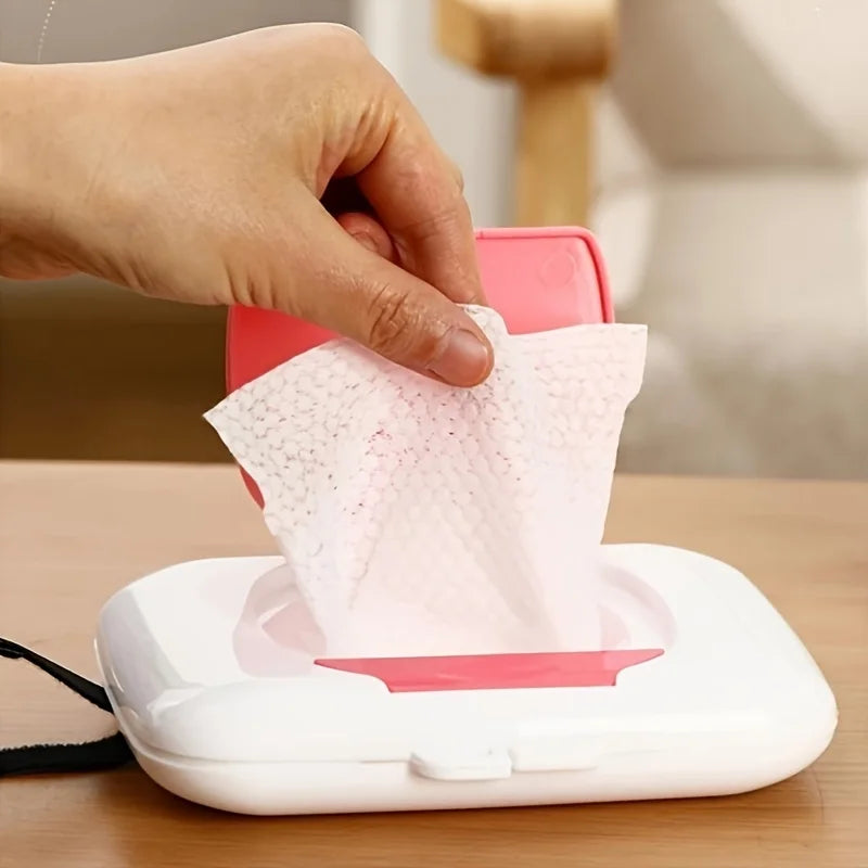 A hand is dispensing pink facial wipes from a white refillable wipe dispenser box.