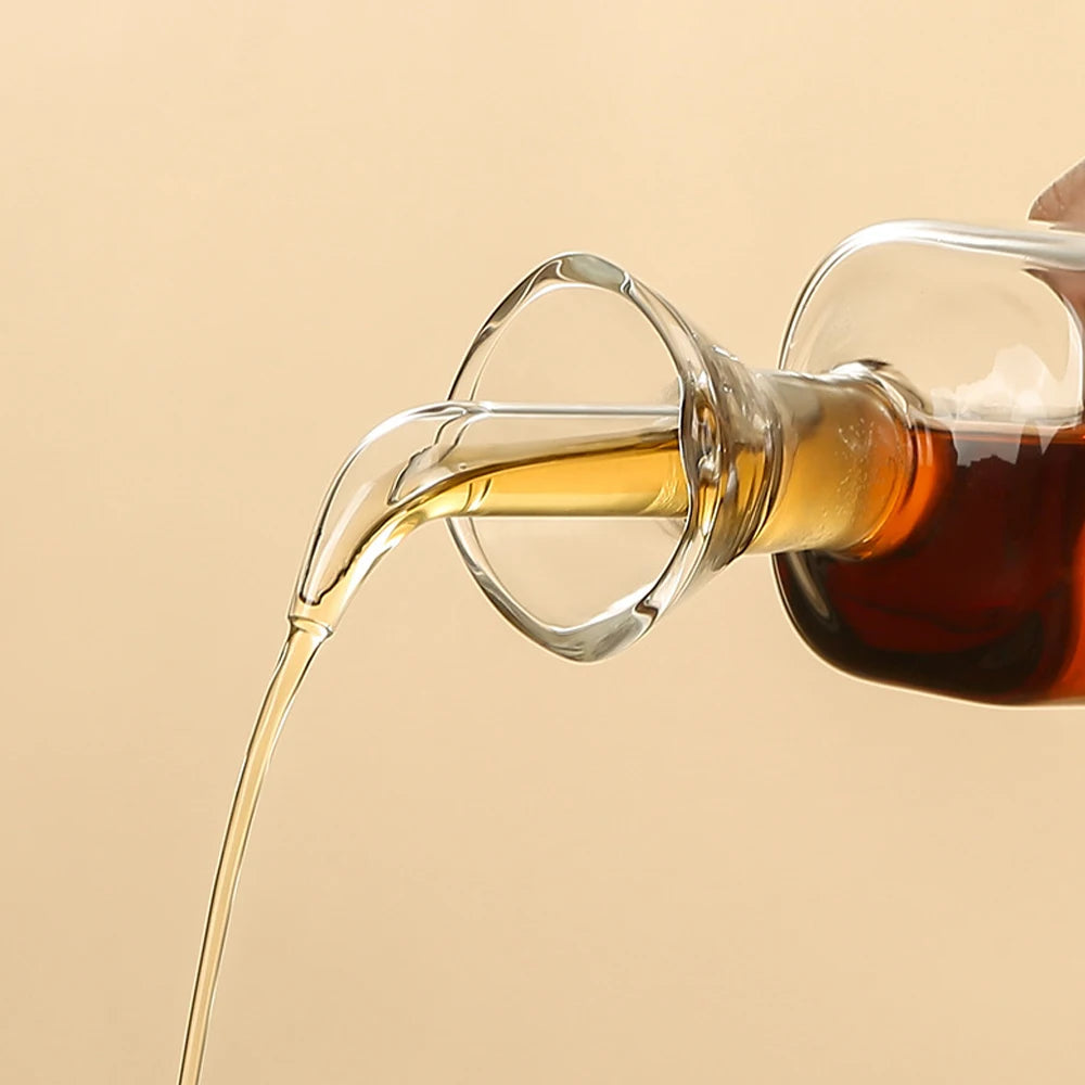 A glass oil and vinegar dispenser is pouring golden liquid into a glass, showing the viscous flow.