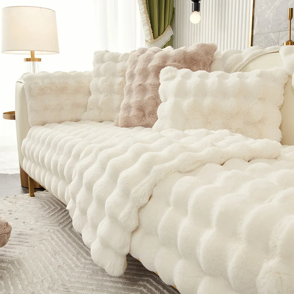 A white fluffy decorative pillow cover is displayed on a sofa, featuring soft textures and light neutral tones.