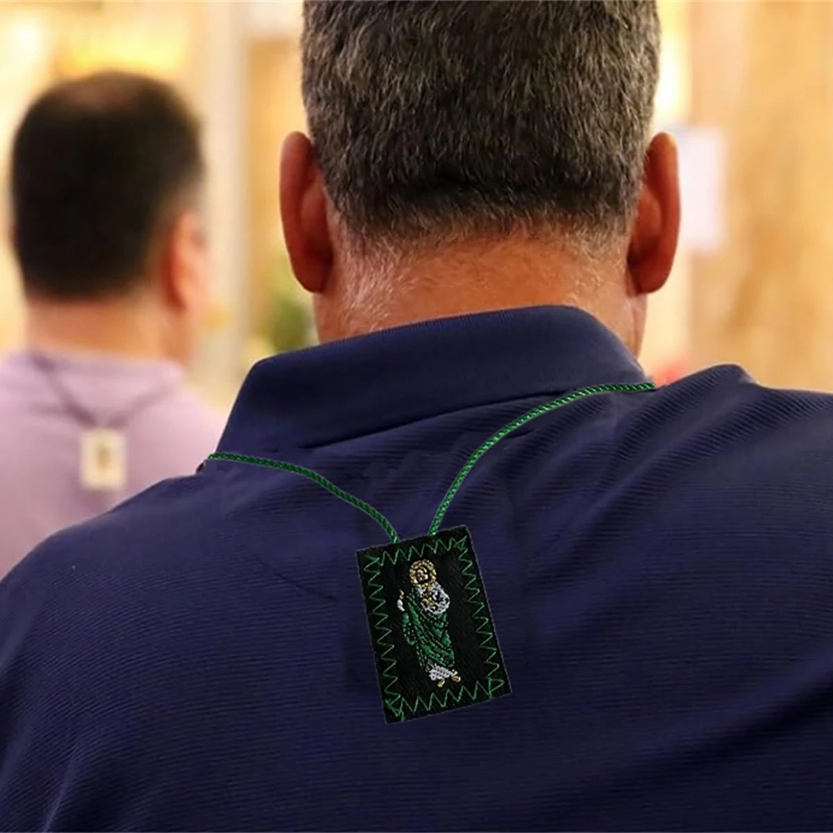 A man wearing a dark blue shirt displays a green scapular necklace with Saint Jude and Our Lady of Guadalupe.