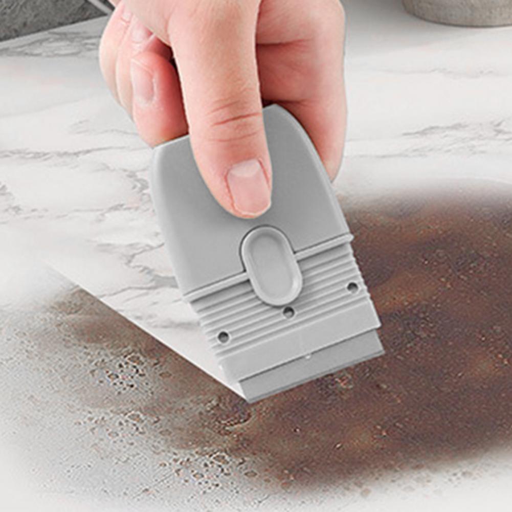 A hand is using a gray double-ended cleaning scraper with a protective cover on a marble surface.