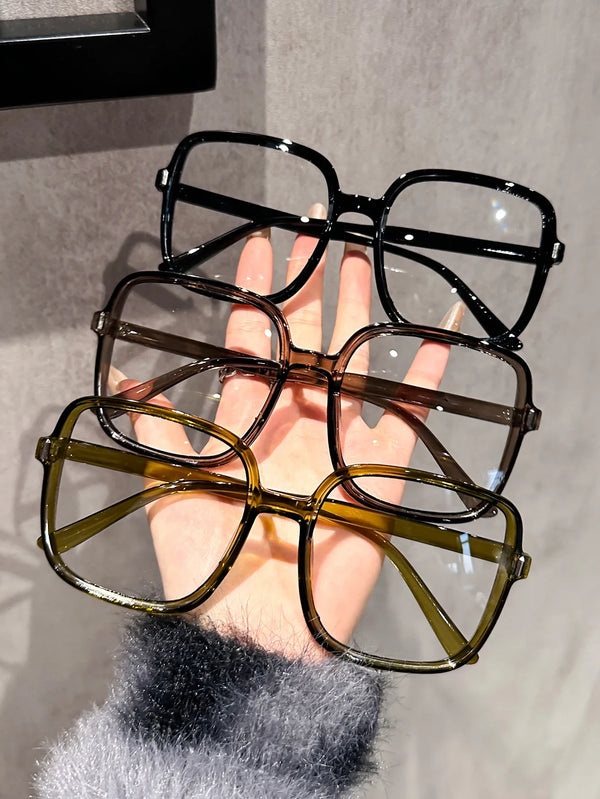 A hand holds a pair of oversized square eyeglasses with a brown frame and clear lenses.
