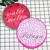 Two circular fruit-themed waterproof shower caps, one red for watermelon and one pink for pitaya, placed on a black grid back