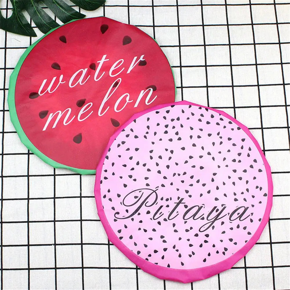 Two circular fruit-themed waterproof shower caps, one red for watermelon and one pink for pitaya, placed on a black grid back