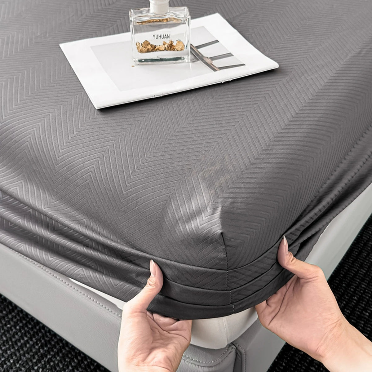 A gray fitted sheet is shown on a bed, with a small glass bottle and scissors placed on top.