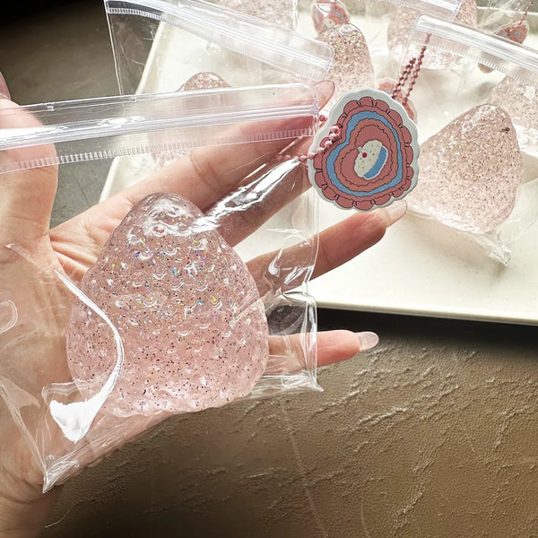 A hand holds a clear plastic bag containing a pink glitter squishy toy and a necklace with a heart pendant.