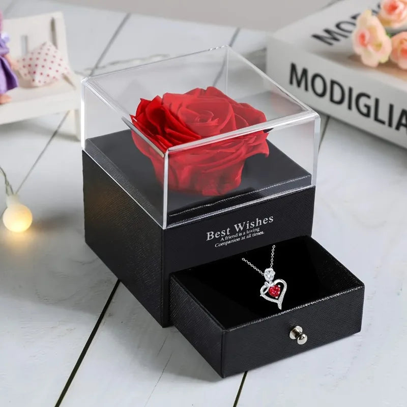 A black box with a glass lid holds a red rose and features a heart pendant necklace.
