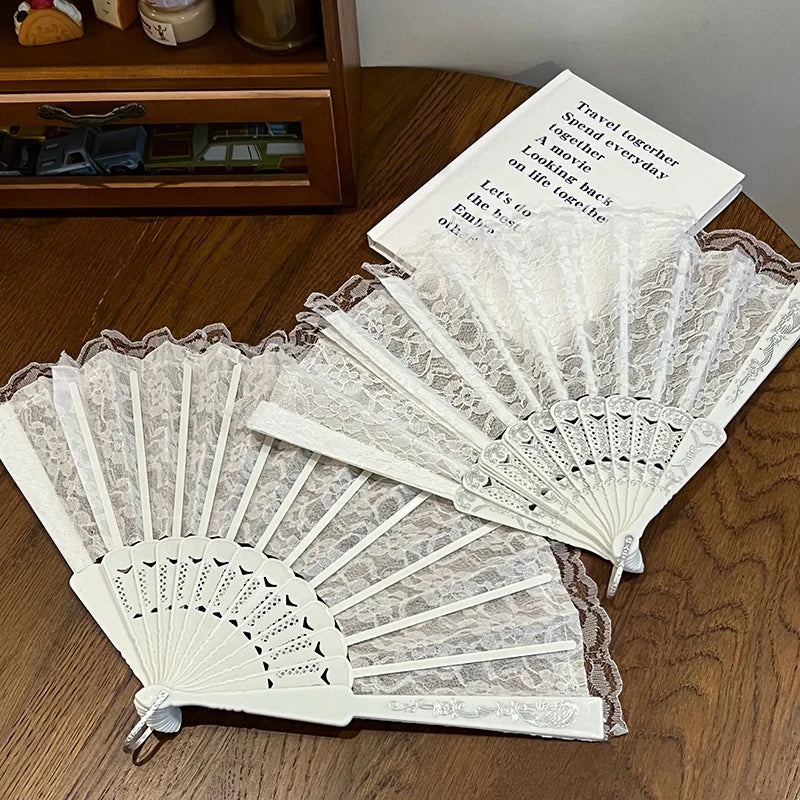 A white vintage lace folding hand fan with a delicate pattern and a brown handle resting on a wooden surface.