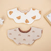 A pair of light beige muslin cotton baby bibs featuring a repeating rainbow pattern and small bear designs.
