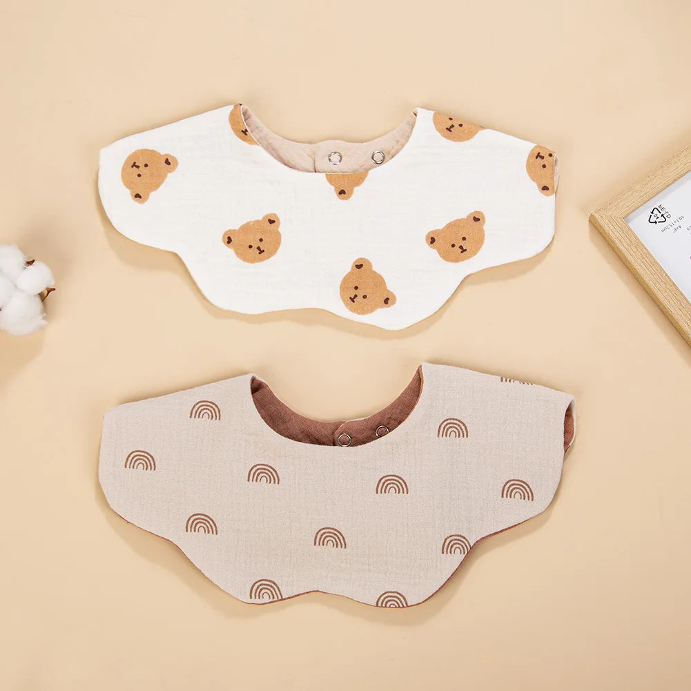 A pair of light beige muslin cotton baby bibs featuring a repeating rainbow pattern and small bear designs.