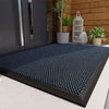 A dark blue non-slip doormat with a diamond pattern placed on a light gray floor near a wooden wall.