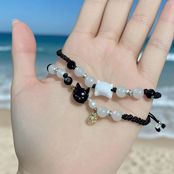 A hand displays a set of minimalist bracelets featuring black obsidian and white moonstone beads against a blue ocean backgro