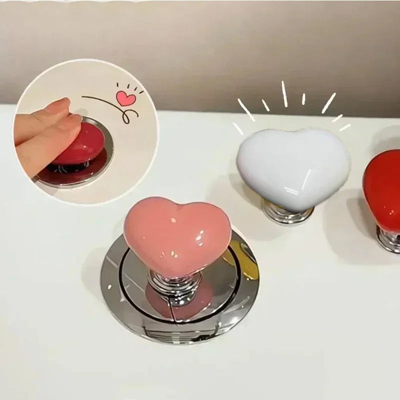A heart-shaped toilet flush button in pink and white is shown on a white surface.
