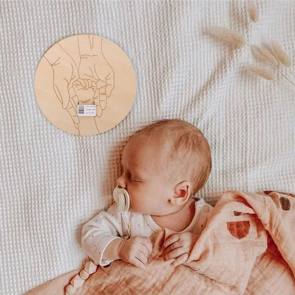 Neugeborenes Baby schläft auf einer weißen Decke neben einem runden Holz-Handabdruck-Anhänger.