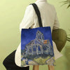 A canvas tote bag featuring a painting of a church building in blue and yellow tones.