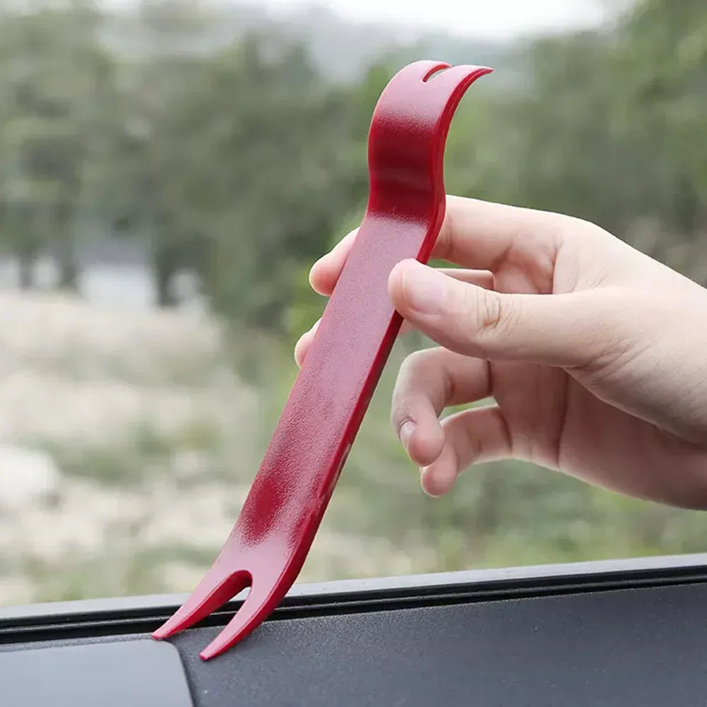 A hand holds a red automotive electrical terminal connector removal tool, showing the tool's shape and size.