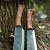A pair of extra-thick manganese steel chopping knives with wooden handles resting against a tree trunk.