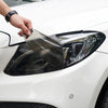 A hand is applying a matte smoke tint vinyl wrap film to a car's headlight assembly.