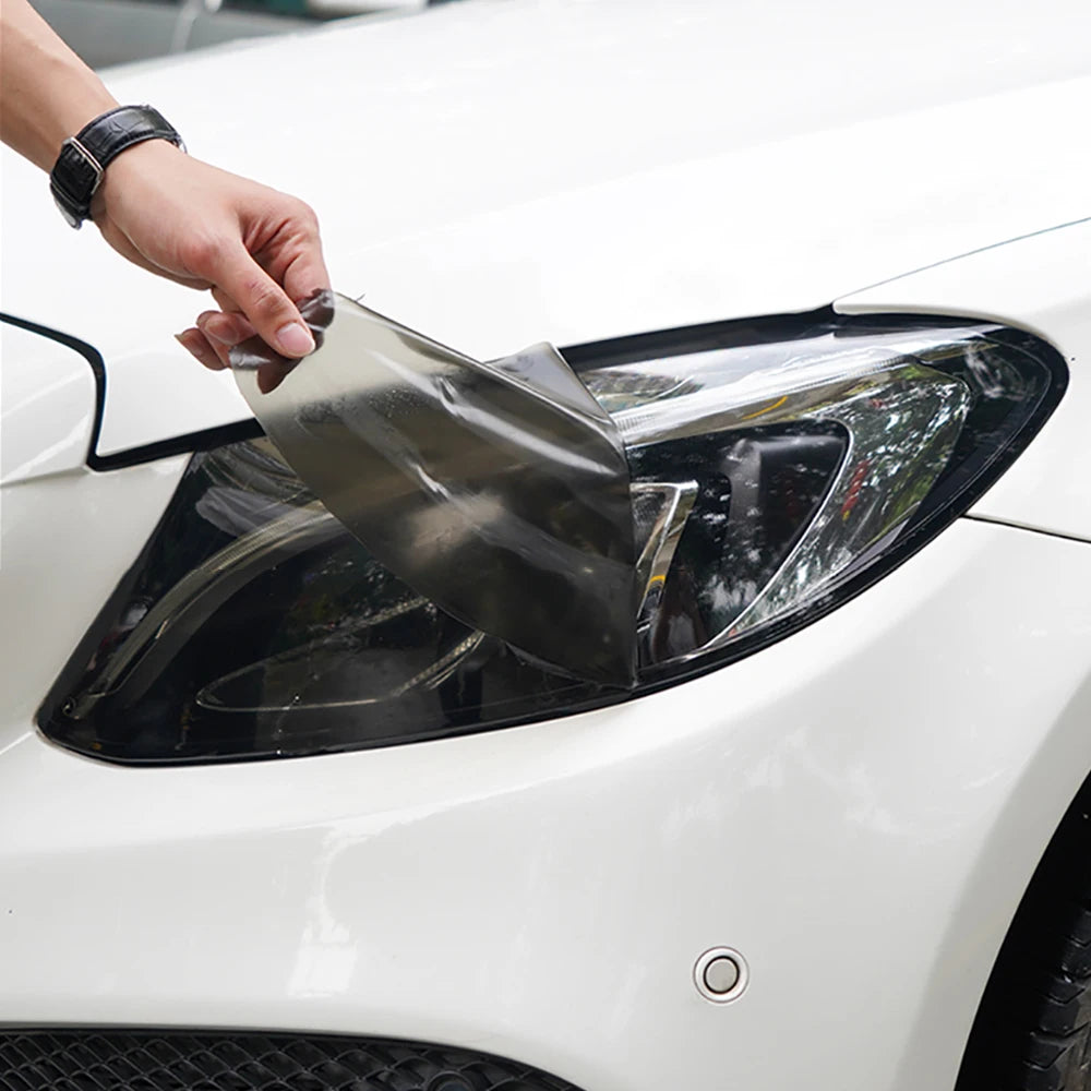 A hand is applying a matte smoke tint vinyl wrap film to a car's headlight assembly.